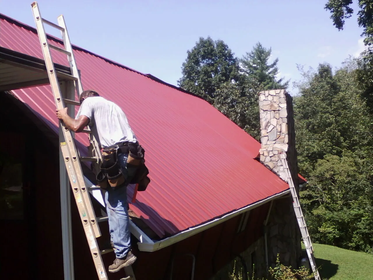 Roofer on a ladder beside a red metal roof and stone chimney — Soffit & Fascia Repair by Roofing Brothers in Morehead